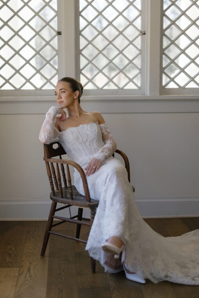 Elegant bridal portrait of a bride in a long sleeve lace gown, seated indoors by a window before her wedding day.