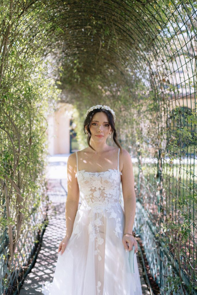 Romantic bridal portrait of a bride in a floral-appliqué dress walking through a vine-covered archway at an outdoor venue.