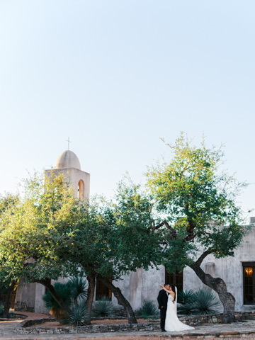 Bride and groom standing in front of chapel and oak trees at Lost Mission, a Central Texas wedding venue with Spanish charm.