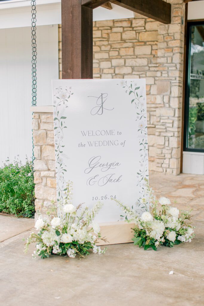 Elegant white wedding welcome sign with greenery border, displayed with white florals in front of a stone venue entrance.