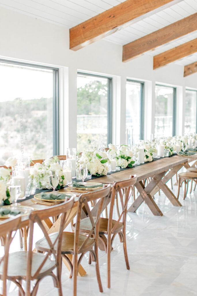 Long wooden wedding reception table set at the Videre Estate with white floral arrangements, gold-rimmed chargers, and glassware in a bright venue with large windows. Using a realistic wedding budget helped bring this together!