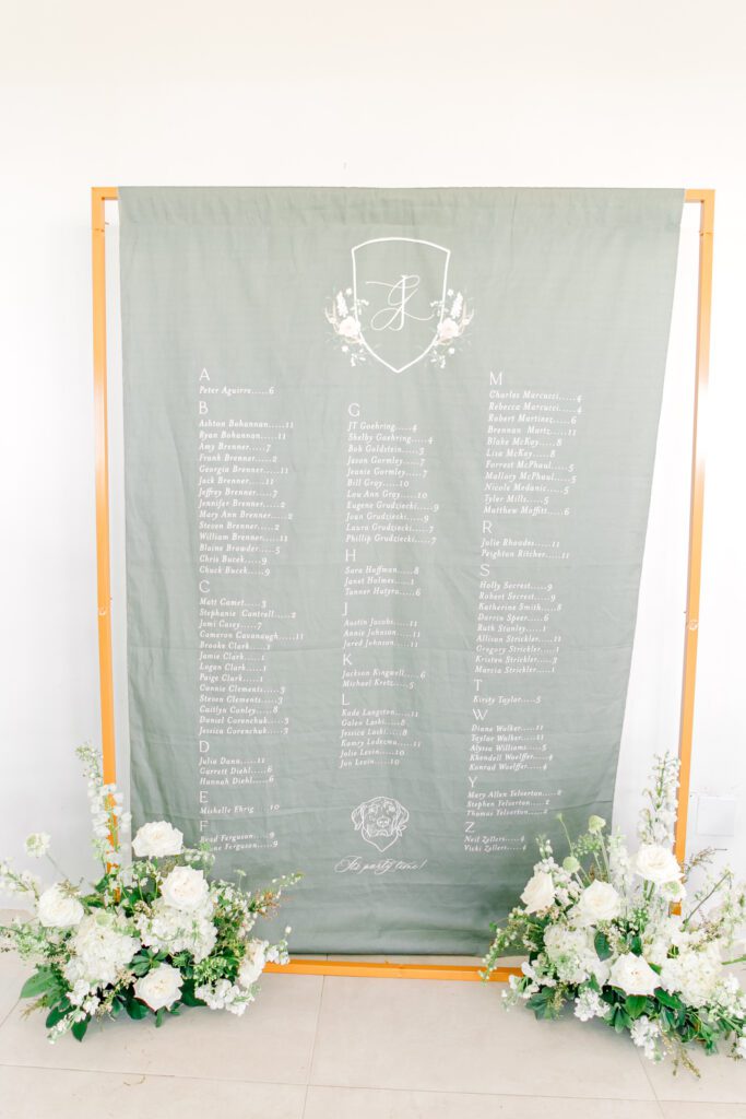 Green fabric wedding seating chart with white calligraphy and floral crest, framed by gold stand and white floral arrangements at the base.