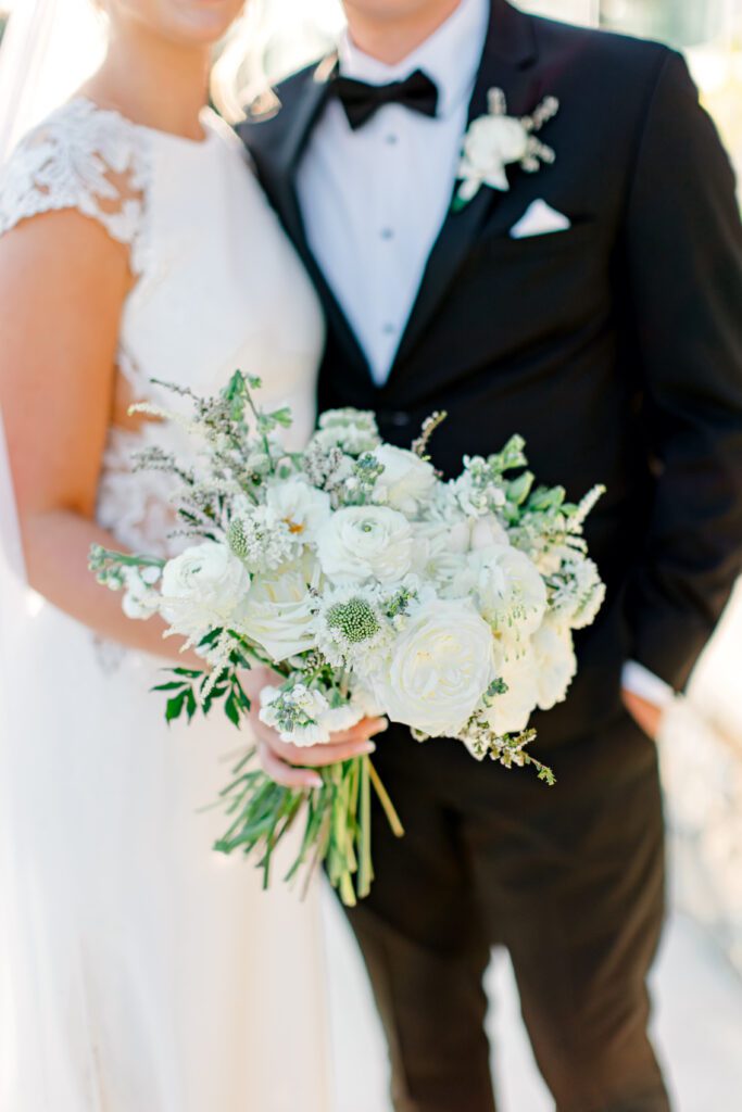 Bride in lace-sleeved wedding gown holding a white rose and ranunculus bouquet, standing beside groom in black tuxedo.