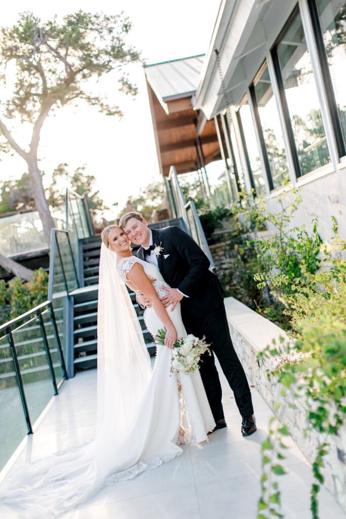 Bride in a fitted lace-sleeve gown and groom in a black tuxedo smiling and embracing on the outdoor staircase at The Videre Estate.