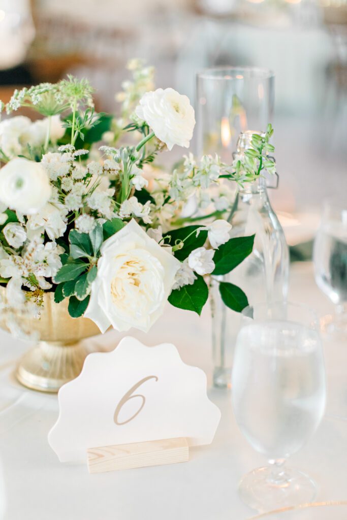 Romantic white and green floral centerpiece in a gold vase with table number and water goblets on a wedding reception table.