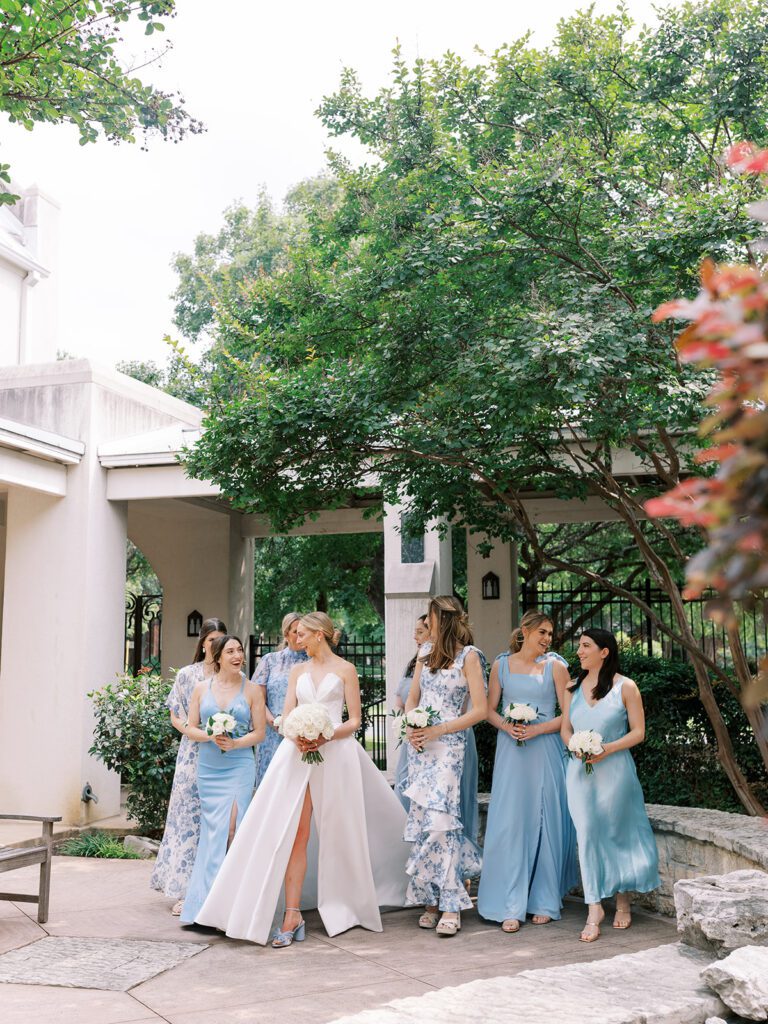 Bride in classic white gown walks with bridesmaids in blue dresses holding white bouquets, illustrating how bridal party size impacts a realistic wedding budget Texas couples should plan for.